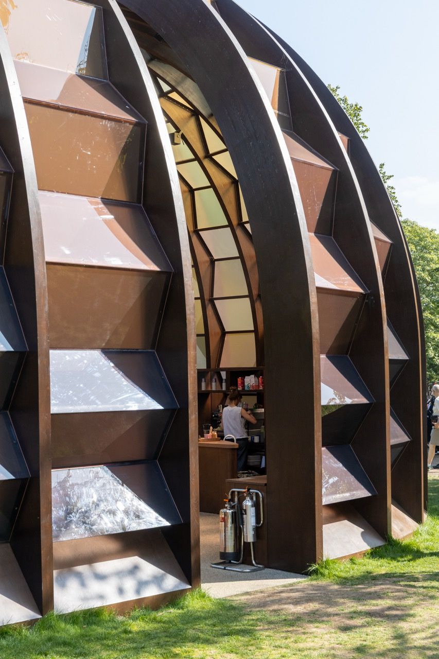 A wide-angle view of the Serpentine Pavilion in a park setting, showing its modern, curved design against a backdrop of trees and a blue sky.