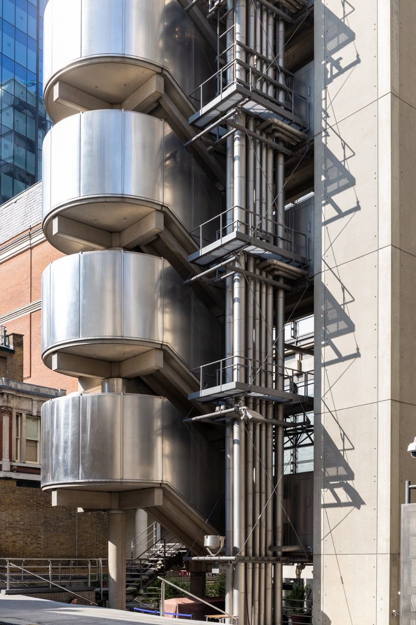A wide-angle, exterior view of the **Lloyd's Building** in London, showcasing its distinctive inside-out architecture with exposed pipes, ducts, and service elements on the facade. The building stands tall against the skyline, reflecting the surrounding urban environment.