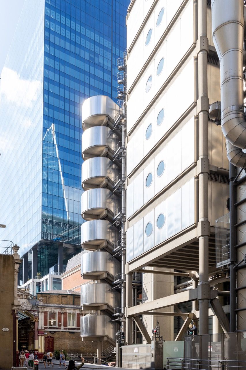 A wide-angle, exterior view of the **Lloyd's Building** in London, showcasing its distinctive inside-out architecture with exposed pipes, ducts, and service elements on the facade. The building stands tall against the skyline, reflecting the surrounding urban environment.
