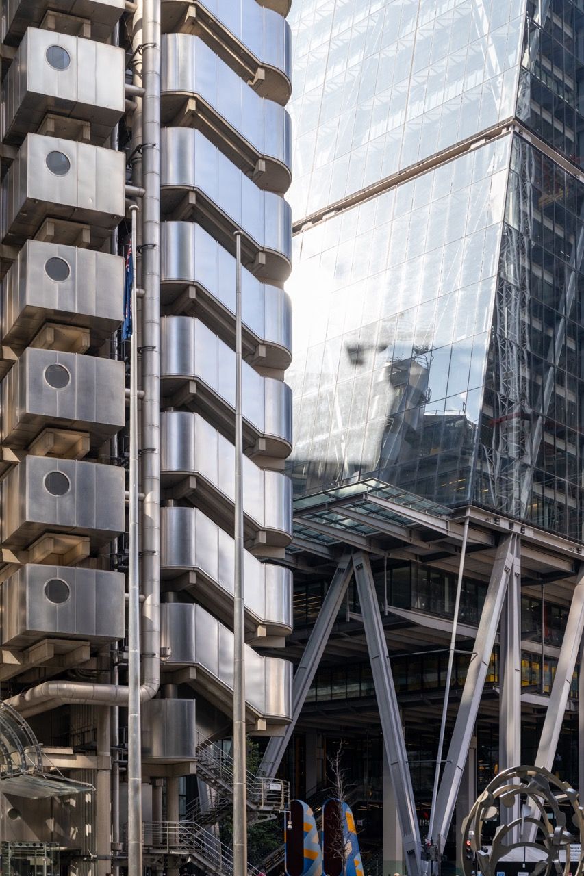 A wide-angle, exterior view of the **Lloyd's Building** in London, showcasing its distinctive inside-out architecture with exposed pipes, ducts, and service elements on the facade. The building stands tall against the skyline, reflecting the surrounding urban environment.