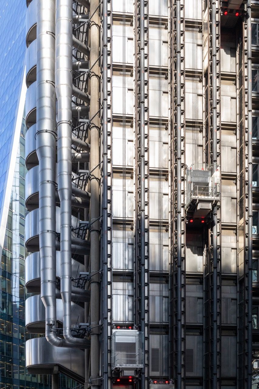 A wide-angle, exterior view of the **Lloyd's Building** in London, showcasing its distinctive inside-out architecture with exposed pipes, ducts, and service elements on the facade. The building stands tall against the skyline, reflecting the surrounding urban environment.