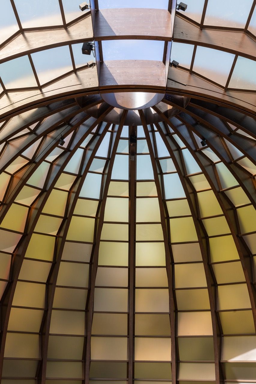 A wide-angle view of the Serpentine Pavilion in a park setting, showing its modern, curved design against a backdrop of trees and a blue sky.