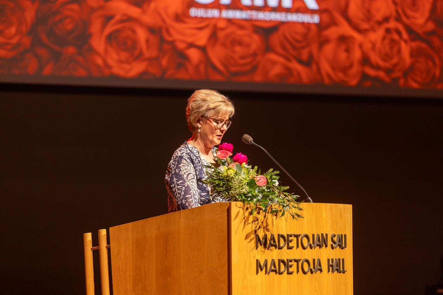 Puhuja Madetojan salin korokkeella, ruusut mikrofonin vieressä.
