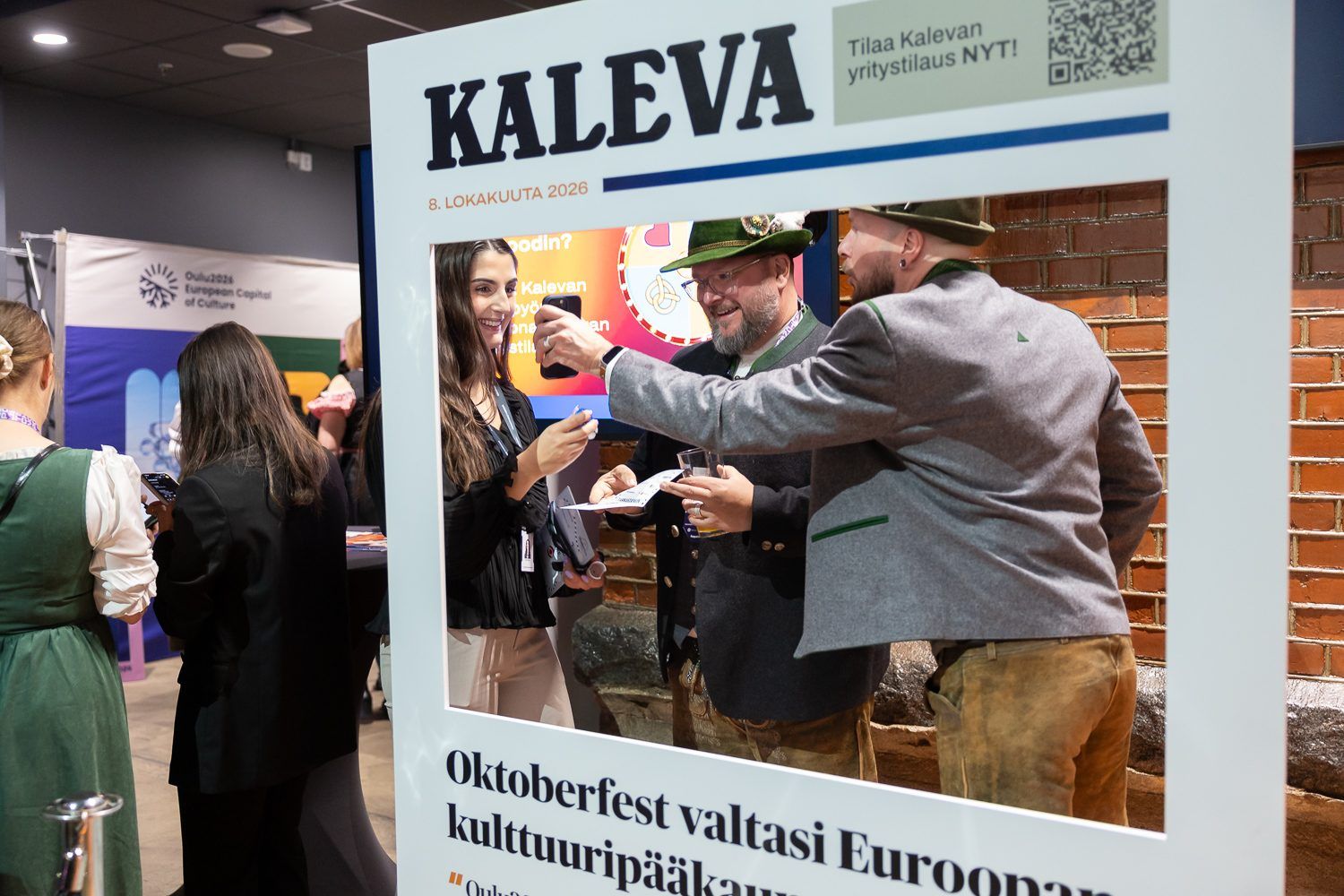 Nainen ja kaksi tirolilaishattuista miestä Kalevan jättikokoisen lehtikehyksen sisällä.