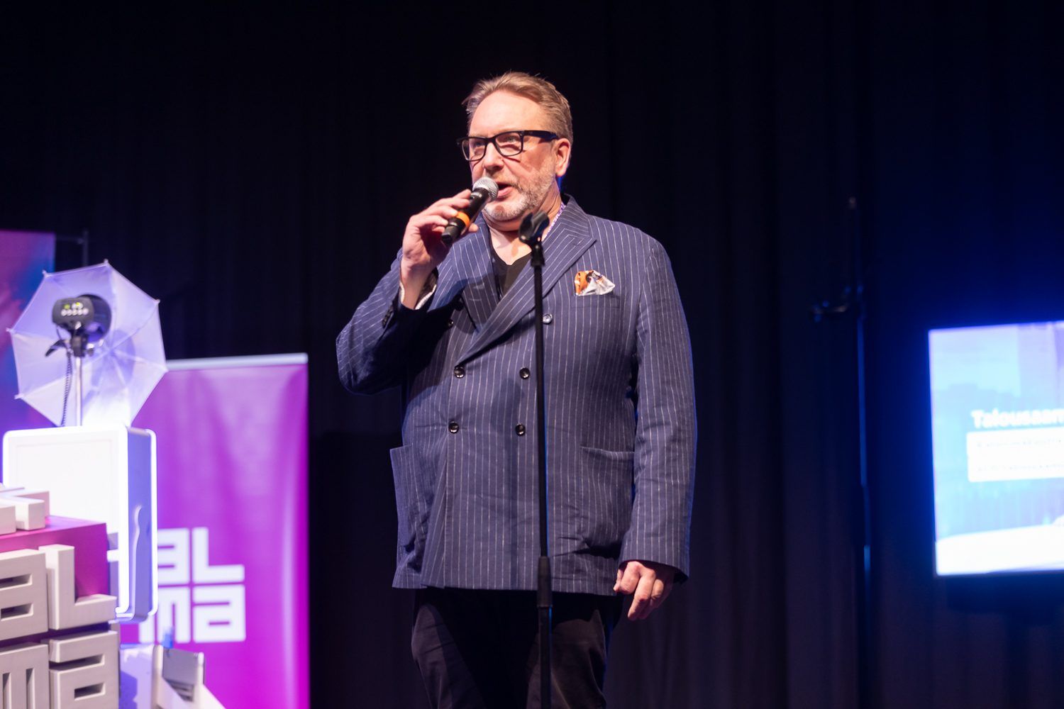 Puhuja sinisessä puvussa puhuu käsimikrofoniin violetin taustaseinän edessä.
