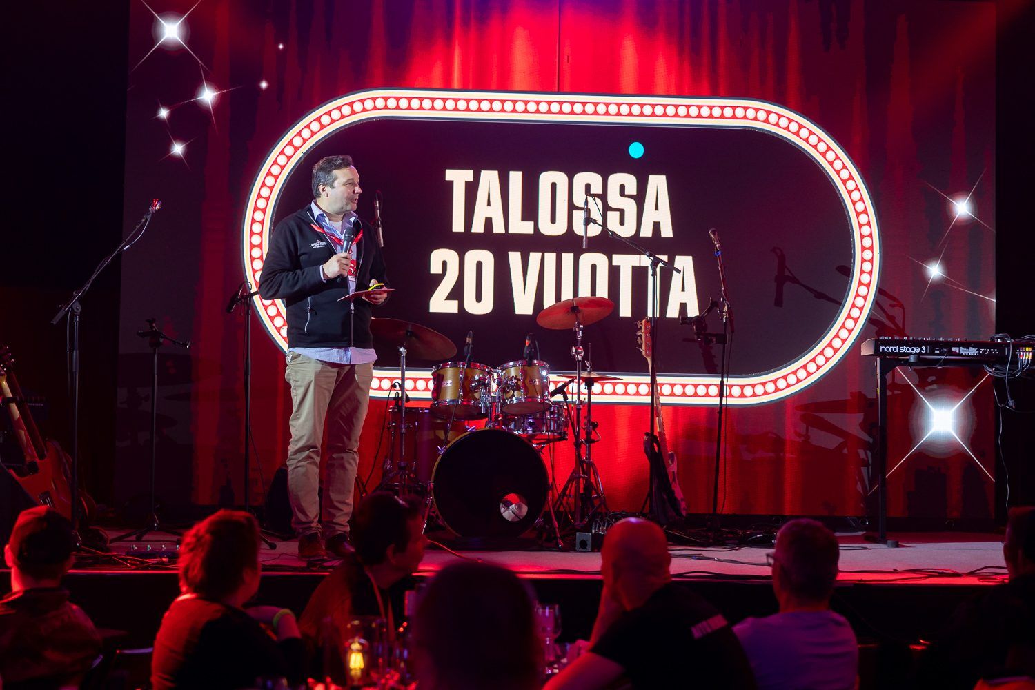 Puhuja lavalla mikrofoni kädessä, takana ruudulla teksti Talossa 20 vuotta.