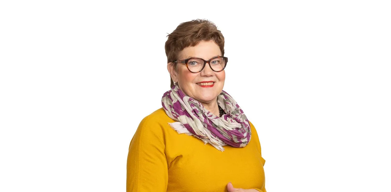 Hallituksen jäsen syvättynä valkoiselle, sinapinkeltainen neule ja kuviohuivi.
