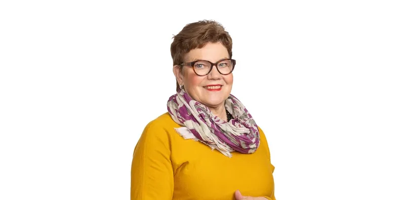 Hallituksen jäsen syvättynä valkoiselle, sinapinkeltainen neule ja kuviohuivi.