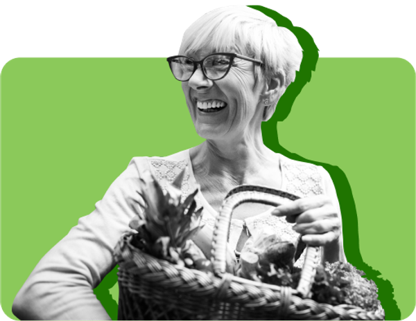 An older woman smiles while holding a basket of groceries against a green background 