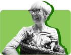 An older woman smiles while holding a basket of groceries against a green background 