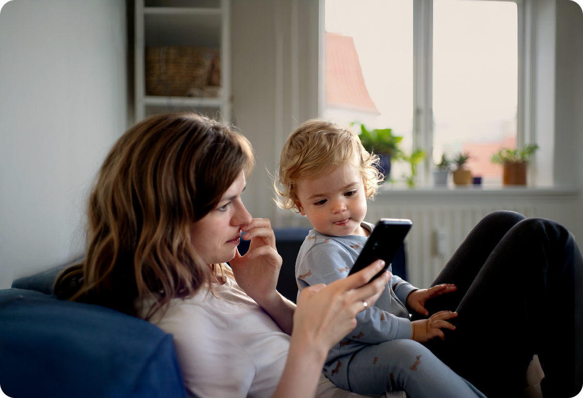 A mom at home, sitting on a sofa with a toddler in her lap, looking at her phone with an interested expression.