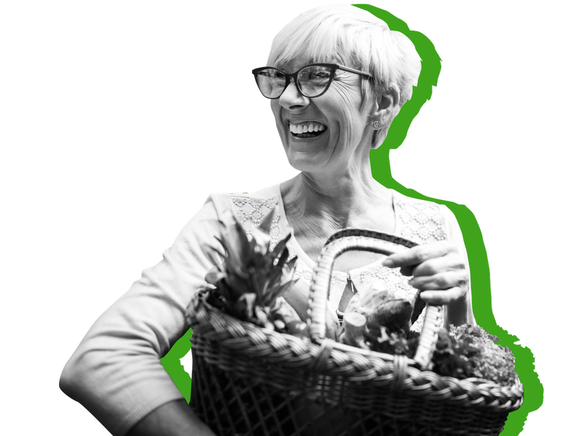 An older woman smiles while holding a basket of groceries against a green background