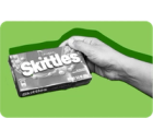 A hand holds up a box of Skittles candy.