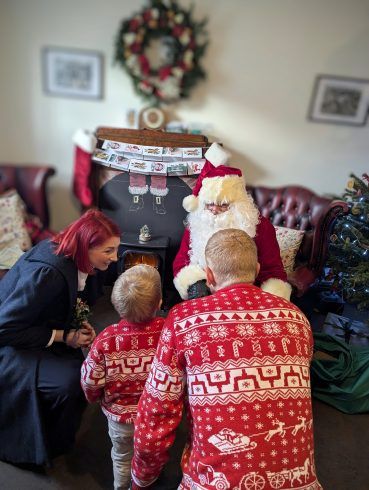 Two children in red Christmas sweaters face a seated Santa beside a decorated fireplace and Christmas tree, while an adult crouches nearby.