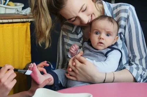 A baby is held by an adult in a striped shirt while another person paints the baby’s foot at a pink table.