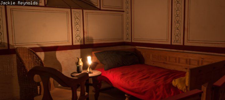 Candlelit reconstructed room with a wooden bed covered by a red blanket, a chair, and two lit lamps on a small table.