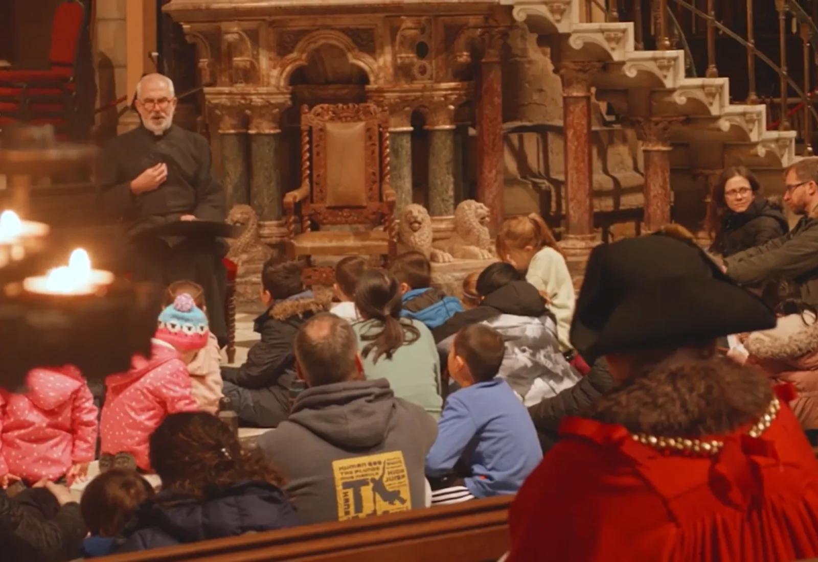 Children and adults sit on the floor inside an ornate cathedral while a robed speaker addresses them; candle flames glow in the foreground.