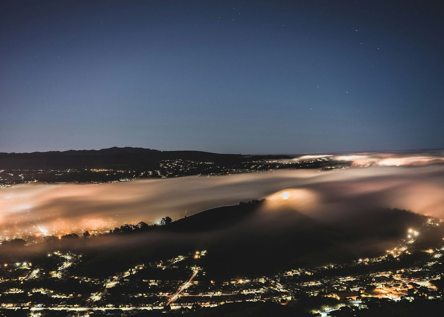 Night photograph of fog filling a valley, with city lights glowing through the mist below a starry sky.