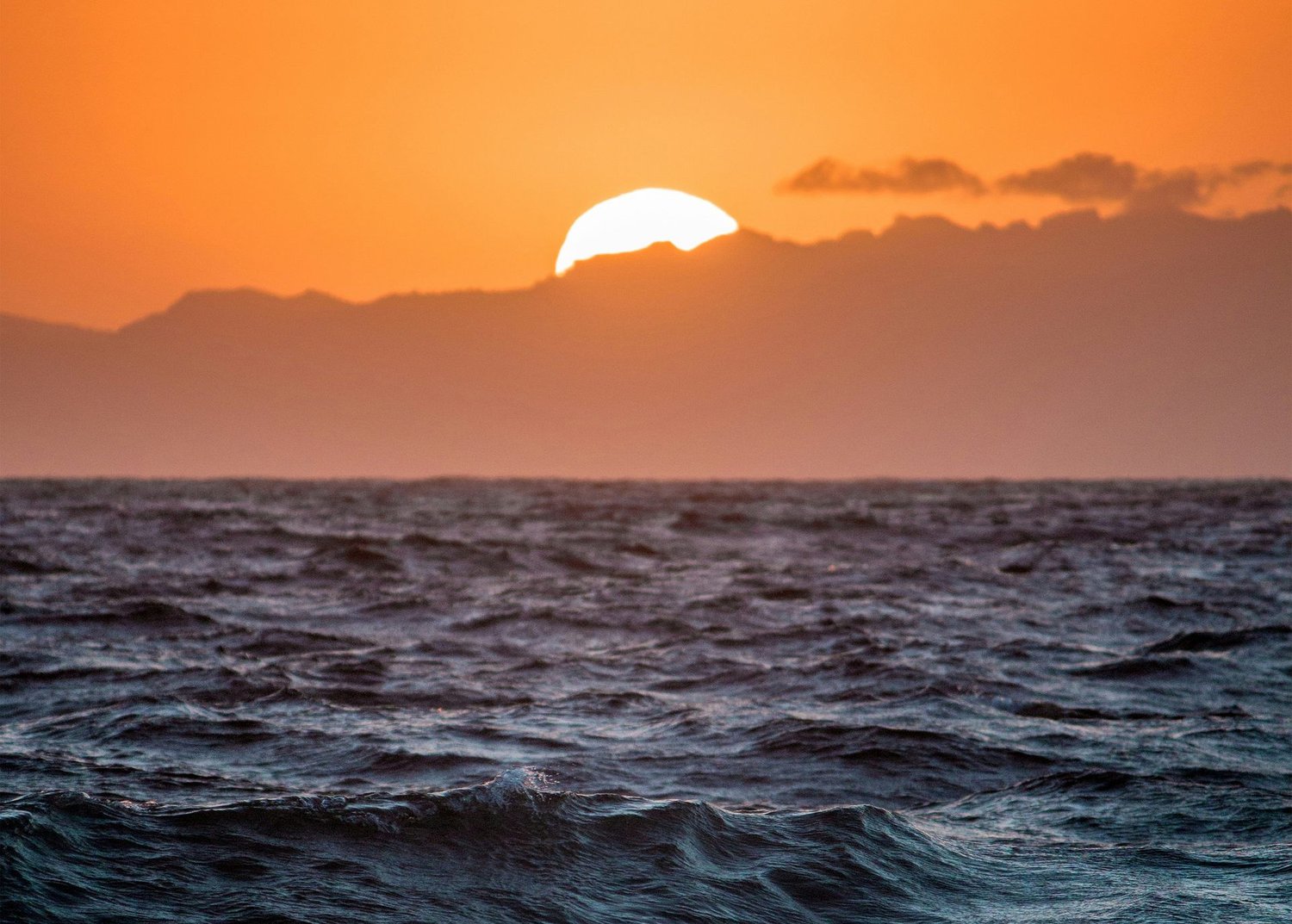 Sunset over dark water, with the sun sitting on a silhouetted mountain range.