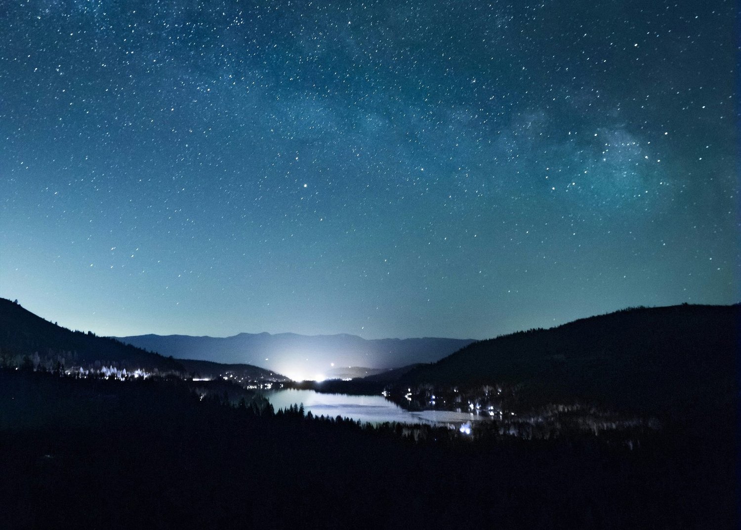 Starry night sky with the Milky Way over a mountain valley, with town lights reflecting on a lake.