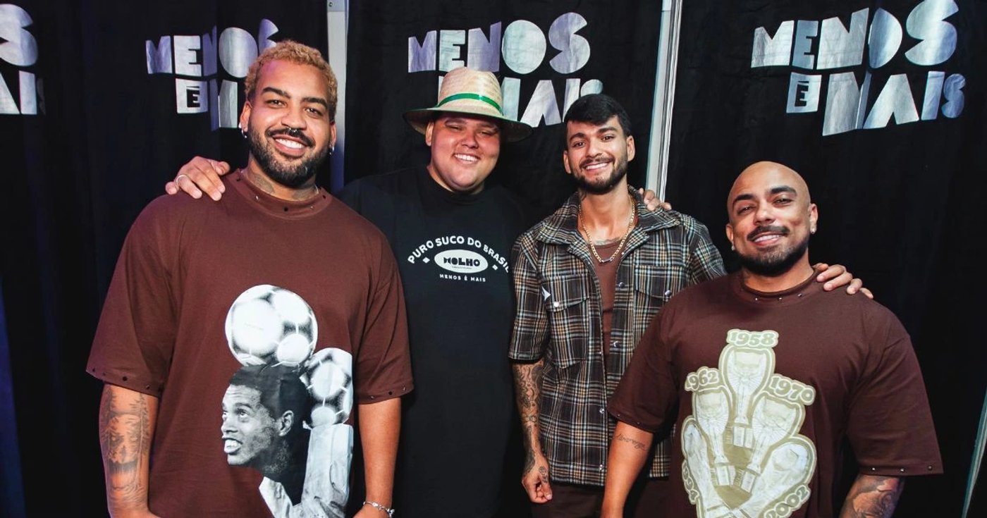 Integrantes do Menos é Mais posam juntos diante de um painel com o nome do grupo.