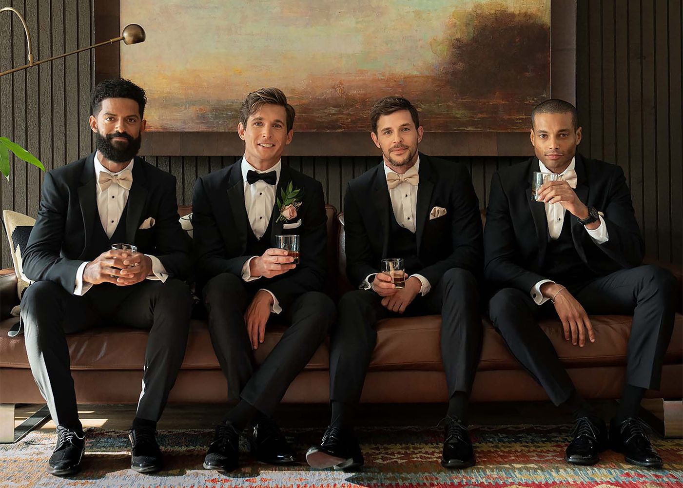 Four men sitting on a couch in black tuxedos 