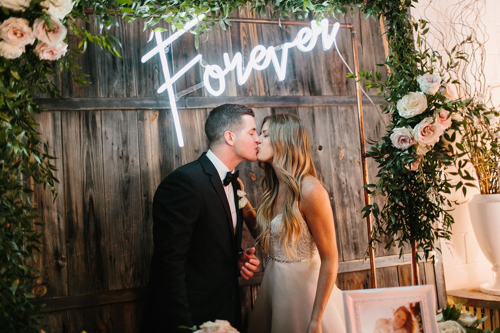photo of a young bride and groom in a black tuxedo kissing under a neon sign that says forever