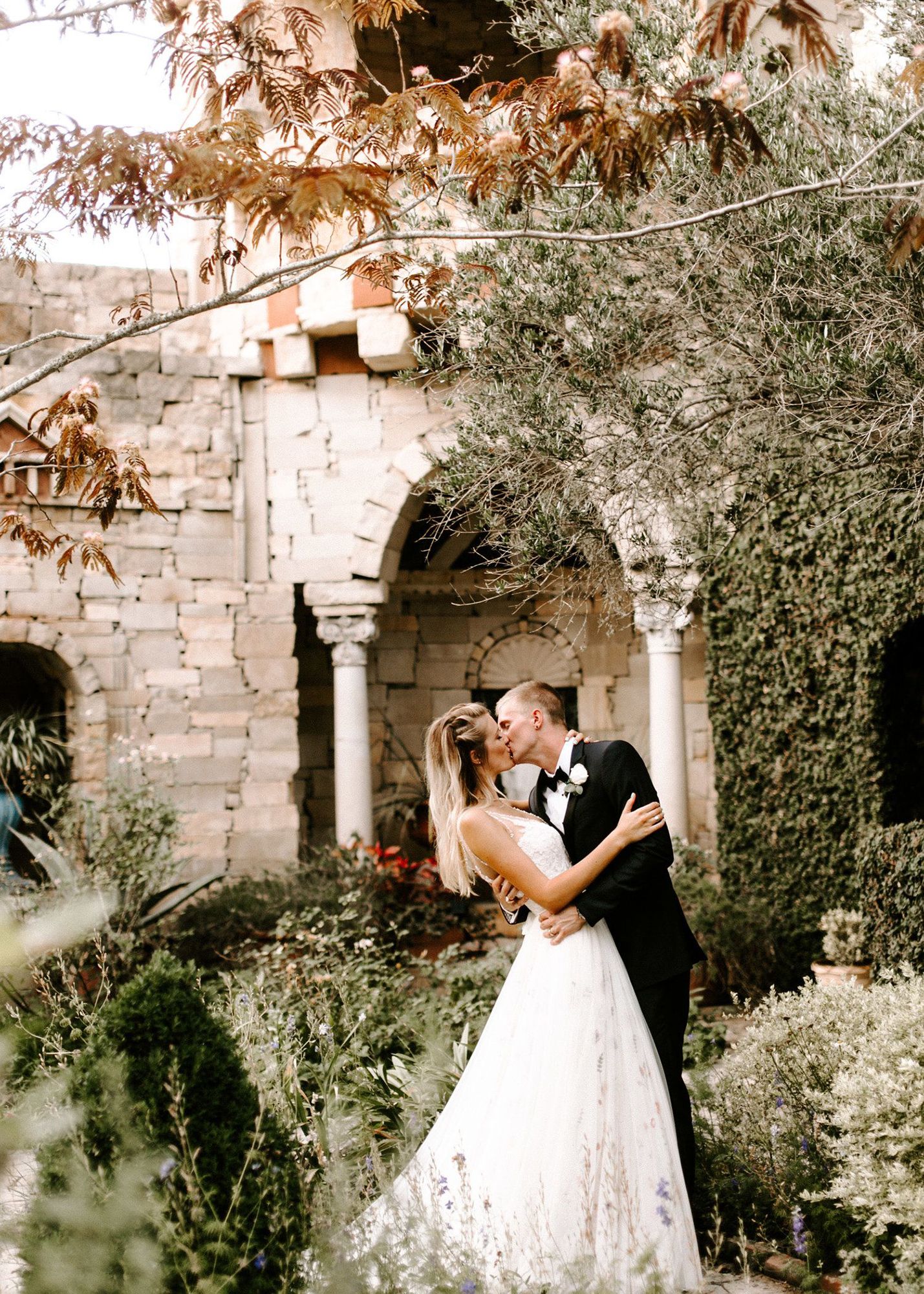 photo of a bride and groom in a black tuxedo kissing while outdoors in a lush garden setting