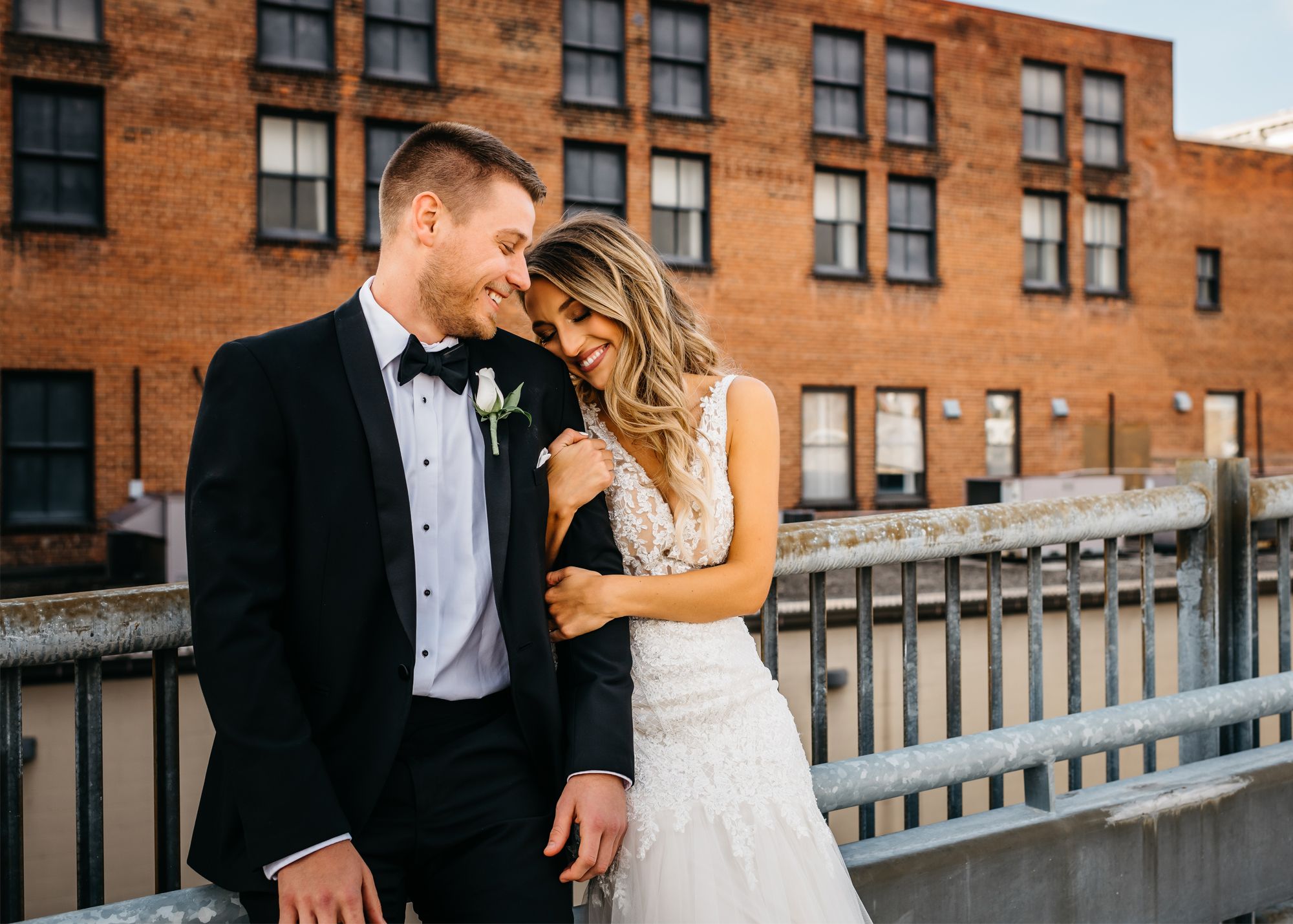 groom in black tuxedo and bride embracing in front of brick building