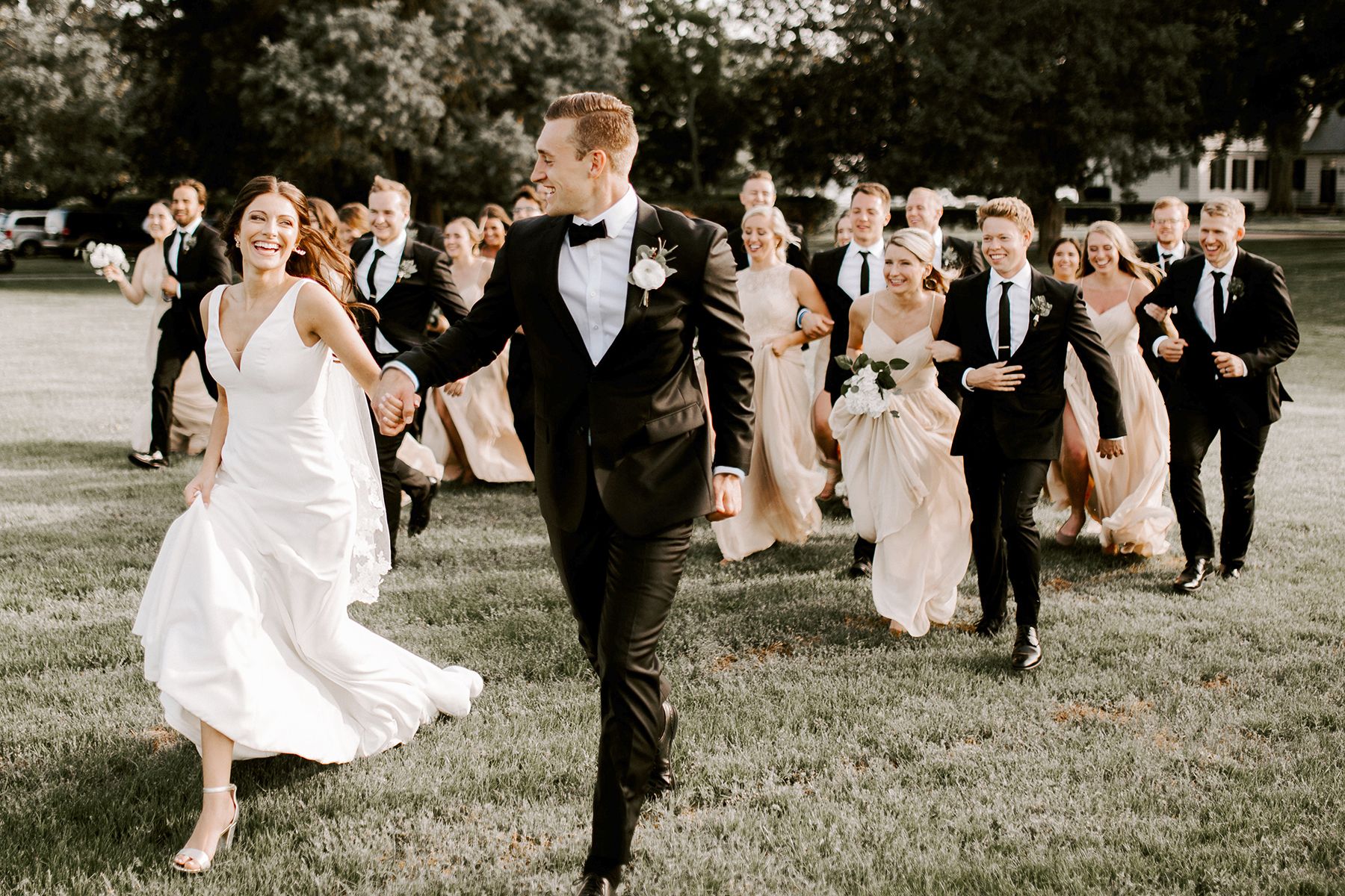 photo of groom in a black tuxedo holding bride’s hand while running outside with wedding party behind