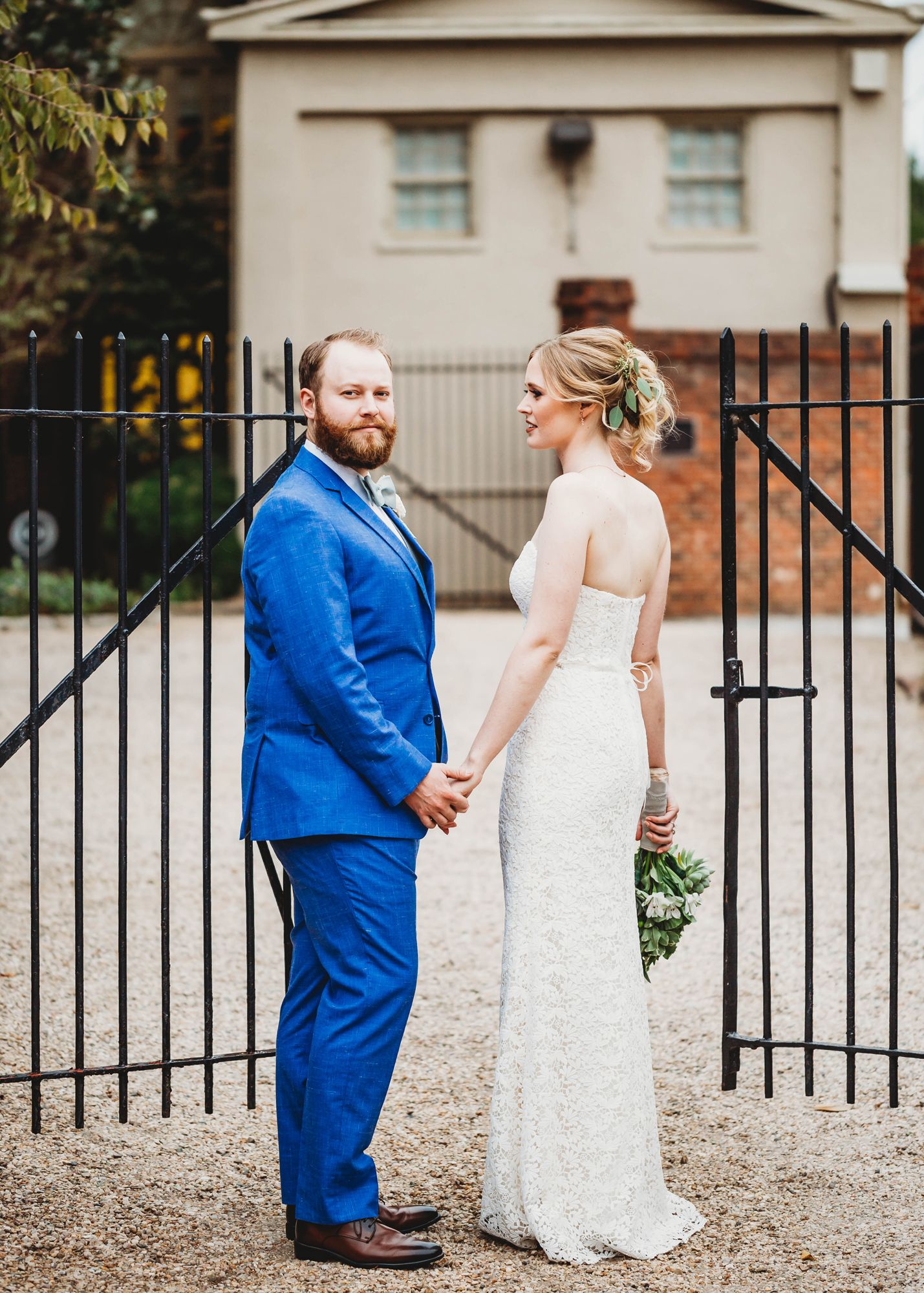 groom in blue suit and bride holding hands in front of wrought iron gate 