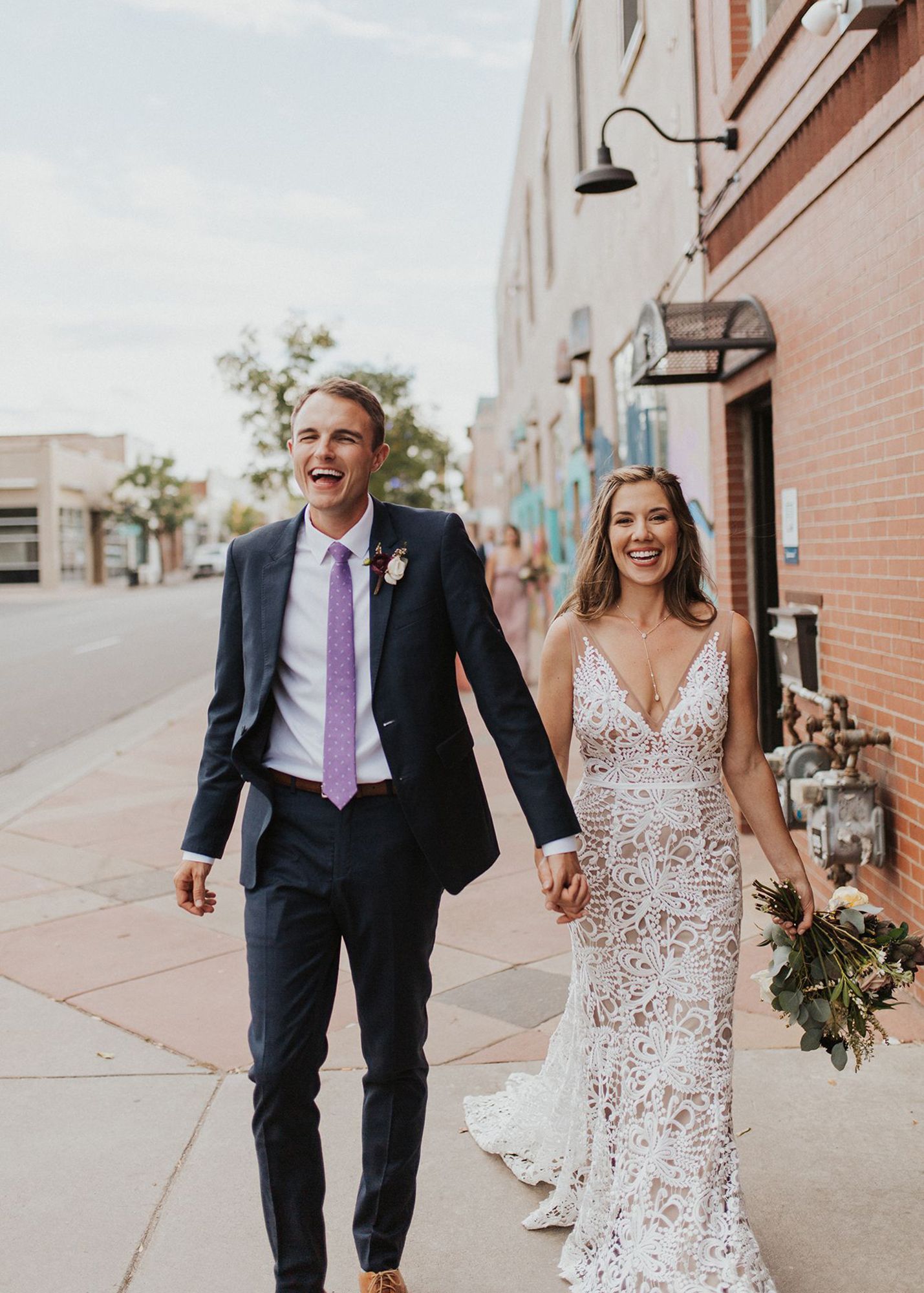  photo of a groom in a blue suit and bride walking outside and smiling while holding hands