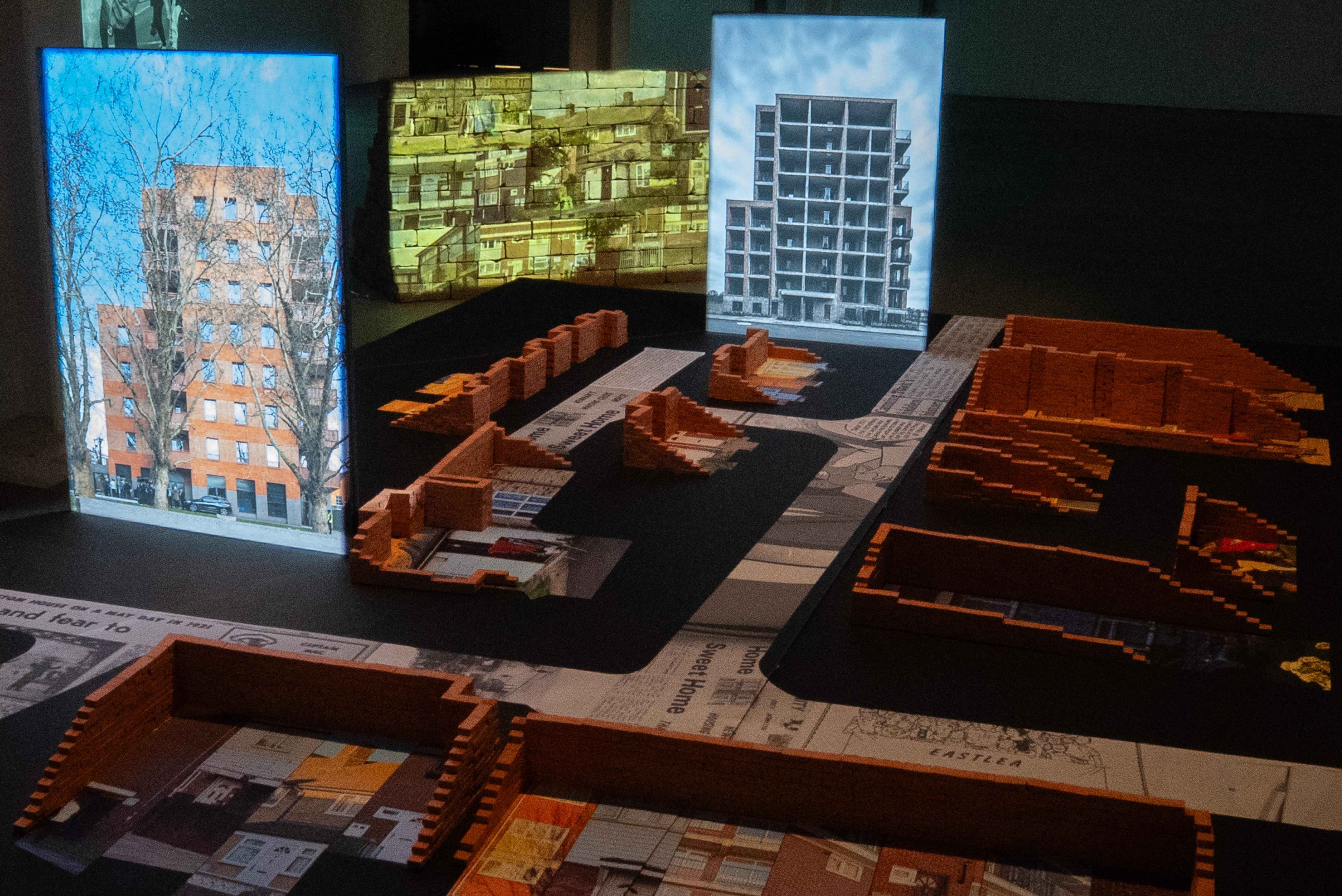 Installation showing brick architectural models representing the remaining buildings of the Custom House Estate, illuminated vertical panels representing the new developments built in place of demolished buildings, and a projection onto a wall of salvaged bricks in the background.