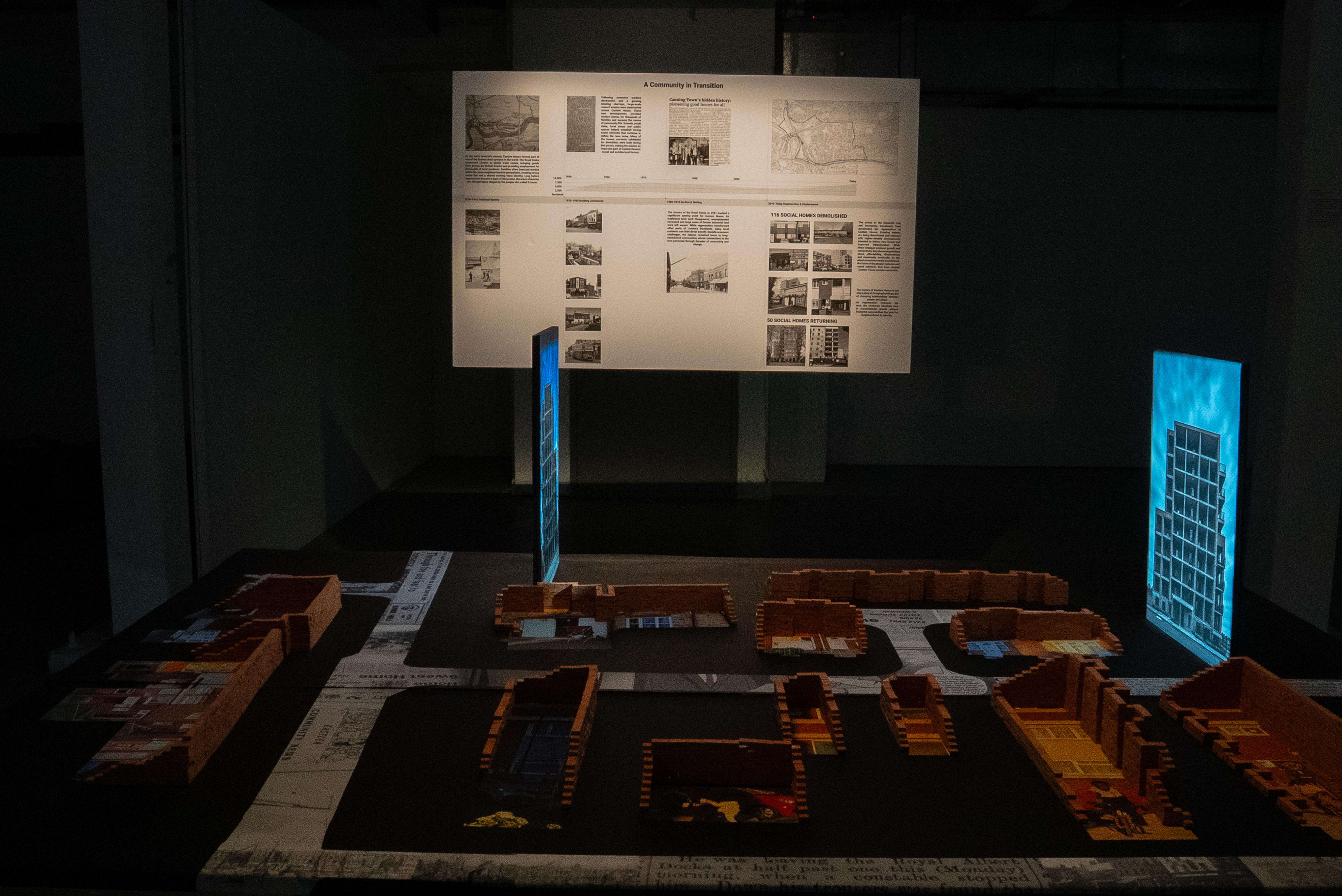 Installation showing brick models of the surviving buildings of the Custom House Estate displayed over archival maps, illuminated panels representing replacement developments, and a timeline documenting the estate's history mounted behind the installation.