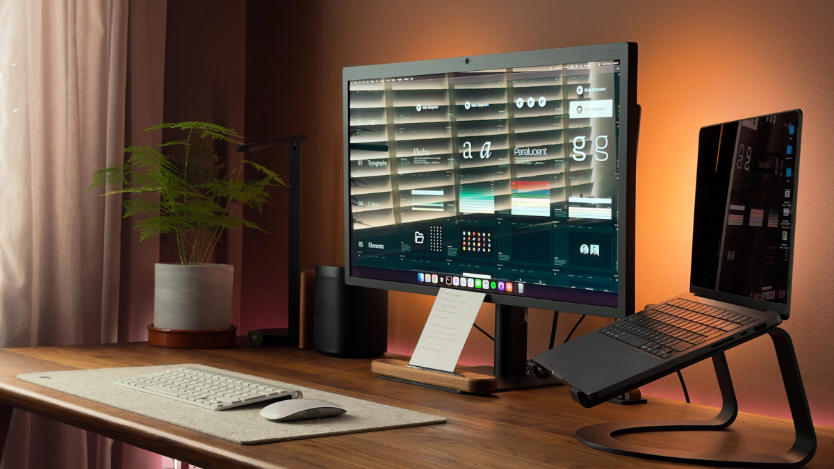 The desk head-on in daylight, with a brand guidelines file on the monitor, a felt desk mat, Magic Keyboard and Mouse, and the wooden to-do stand holding a paper list upright.