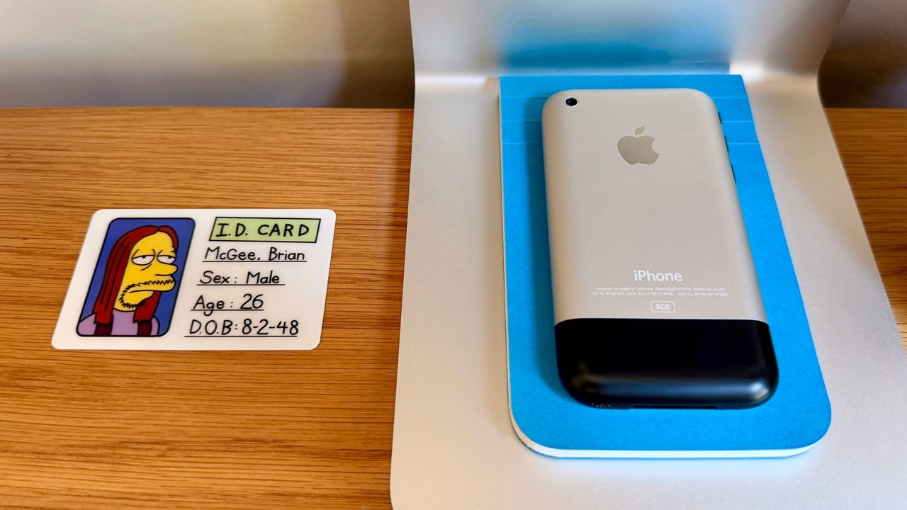 A Simpsons I.D. card for Brian McGee beside an original iPhone displayed in its box.