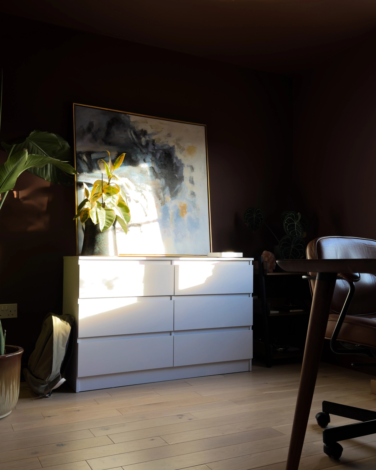 The white drawer unit against the brown wall, with an abstract painting leaning on top and afternoon light raking across the fronts.