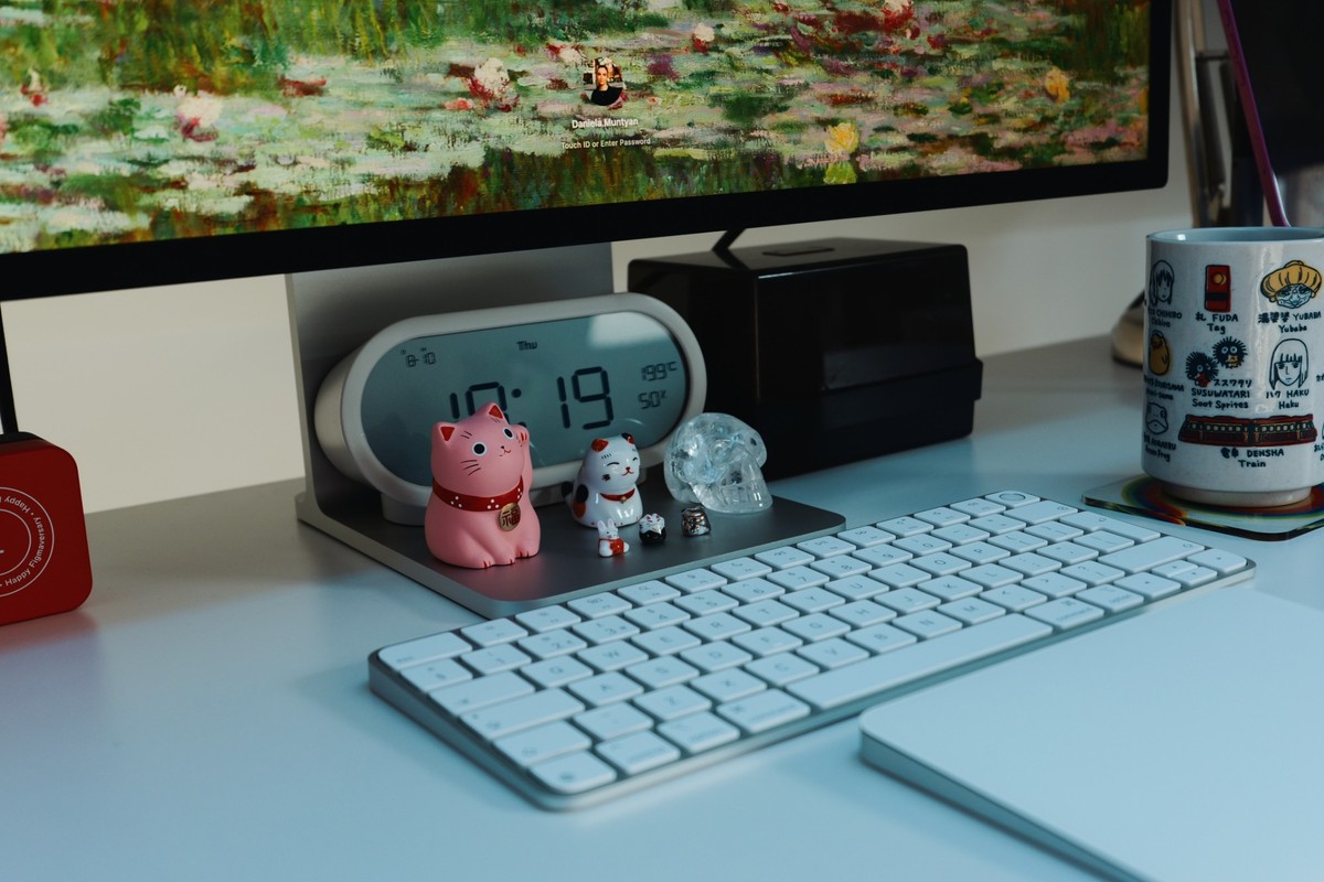 A close view of the monitor stand with a pink and a white lucky cat, a glass skull and a digital clock reading 10:19, beside a Magic Keyboard and a Ghibli mug.