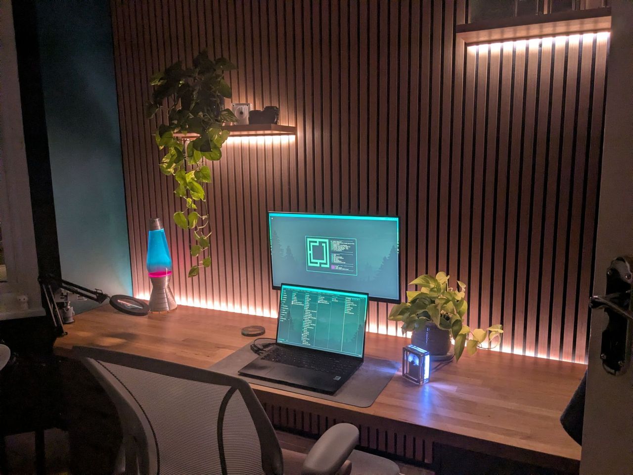 The desk at night with the slatted wall backlit in warm orange, a blue and pink lava lamp glowing, and teal code on the monitor and laptop.