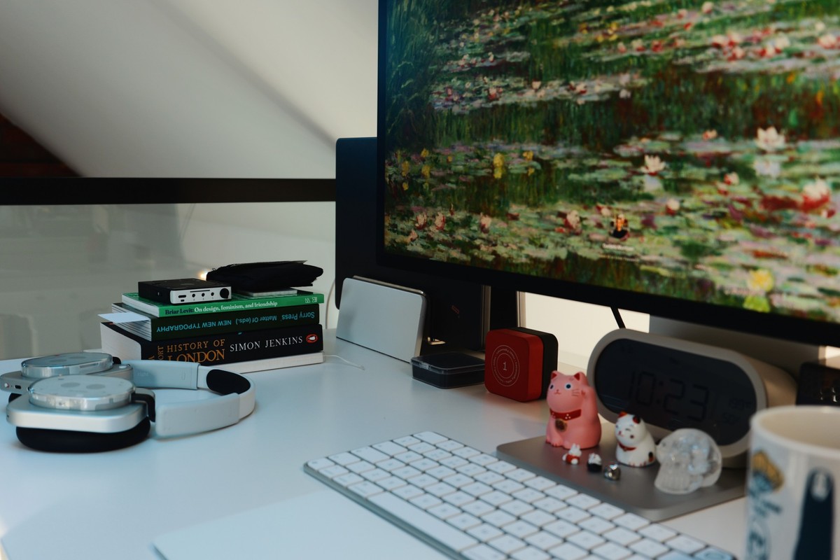 The desk from the side with silver headphones, a desktop DAC on a short stack of books, the Studio Display, a digital clock and two lucky cats beside the keyboard.
