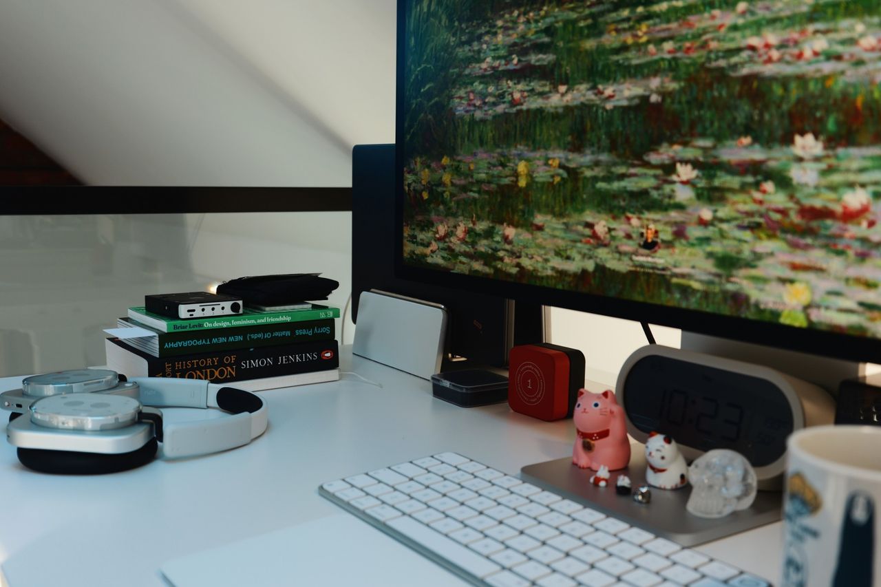 The desk from the side with silver headphones, a desktop DAC on a short stack of books, the Studio Display, a digital clock and two lucky cats beside the keyboard.