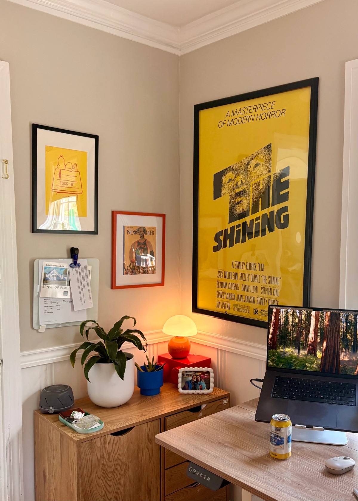 A corner of Zack Davenport's setup with a framed yellow "The Shining" poster above a monitor riser holding plants, a white dome lamp, an orange ball lamp, and a MacBook showing a redwood-forest wallpaper.