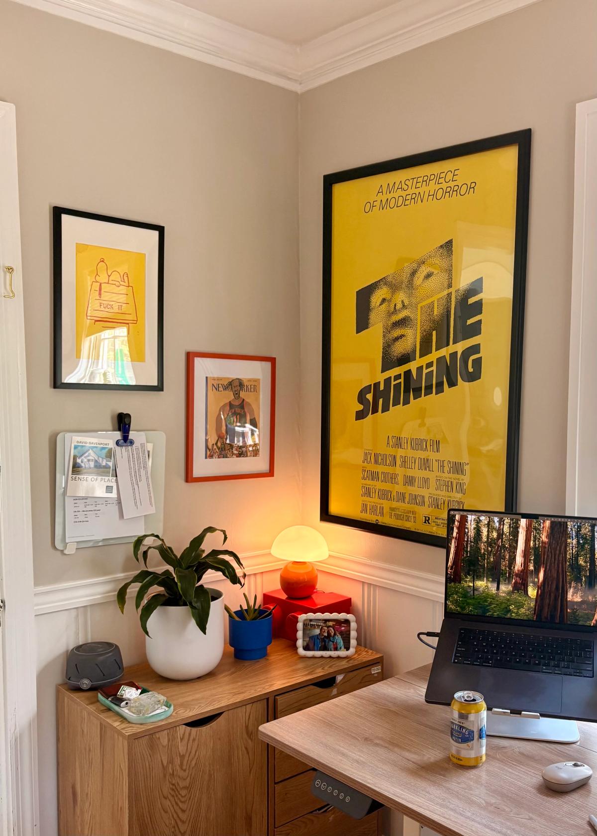 A corner of Zack Davenport's setup with a framed yellow "The Shining" poster above a monitor riser holding plants, a white dome lamp, an orange ball lamp, and a MacBook showing a redwood-forest wallpaper.
