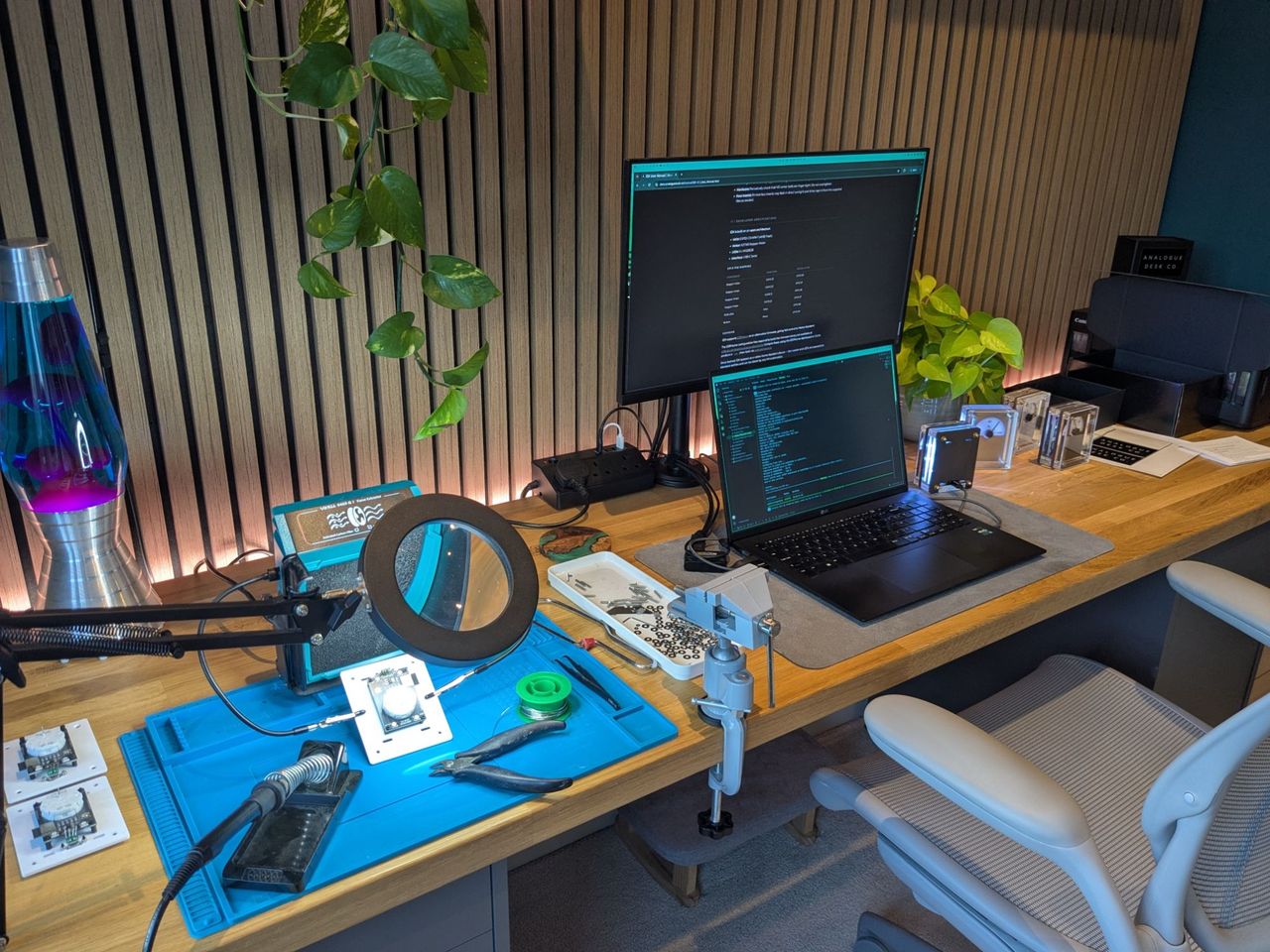 The desk in full production-line mode, with the soldering station and magnifier on the left and the laptop and monitor in the middle.