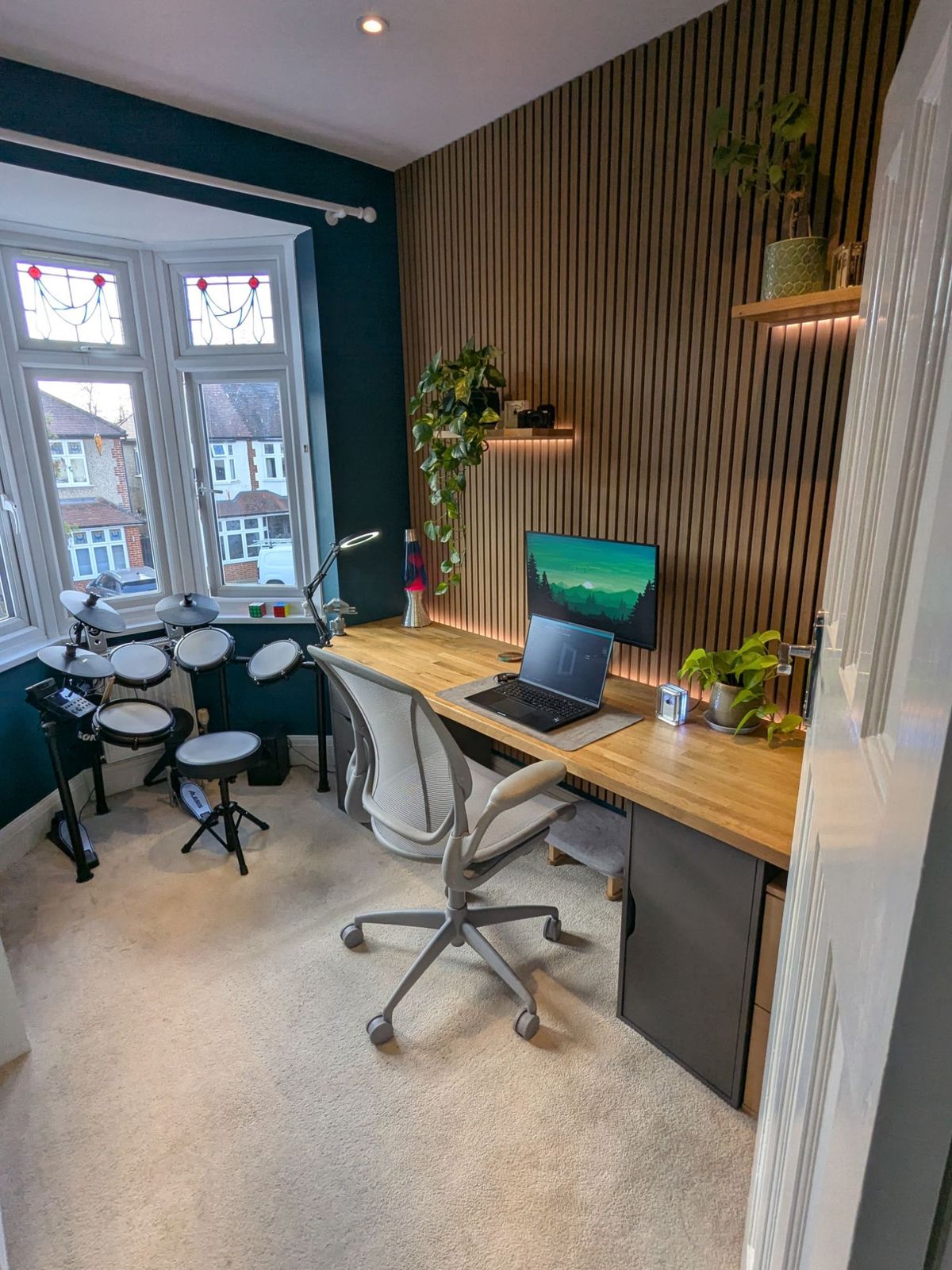 The office from the doorway: the electronic drum kit in the bay window, the oak desk along the slatted wall and a mesh task chair.