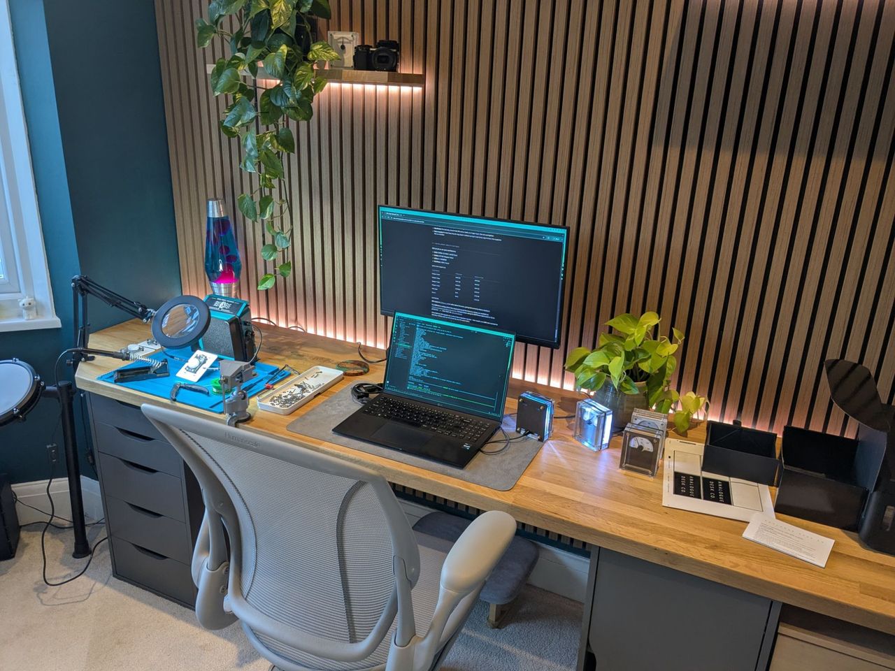 The same desk in workshop mode, with a blue soldering mat, a ring magnifier, a bench vise, a parts tray and a laptop showing code.