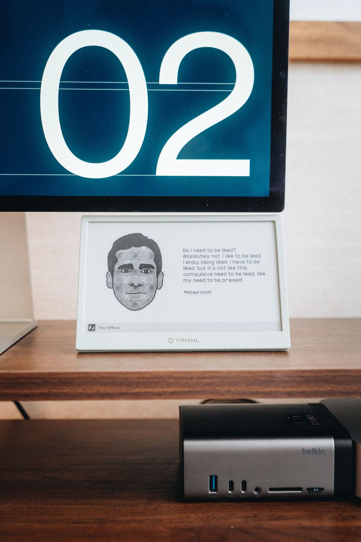E-ink display showing a portrait and a quote on a desk shelf below an Apple Studio Display, with a USB-C hub underneath