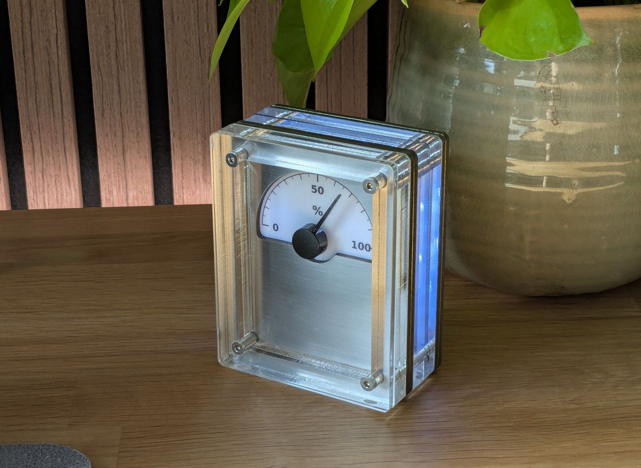 A close view of the IDX desk gauge: a needle dial reading 0 to 100 percent in a clear acrylic case, lit blue from inside.