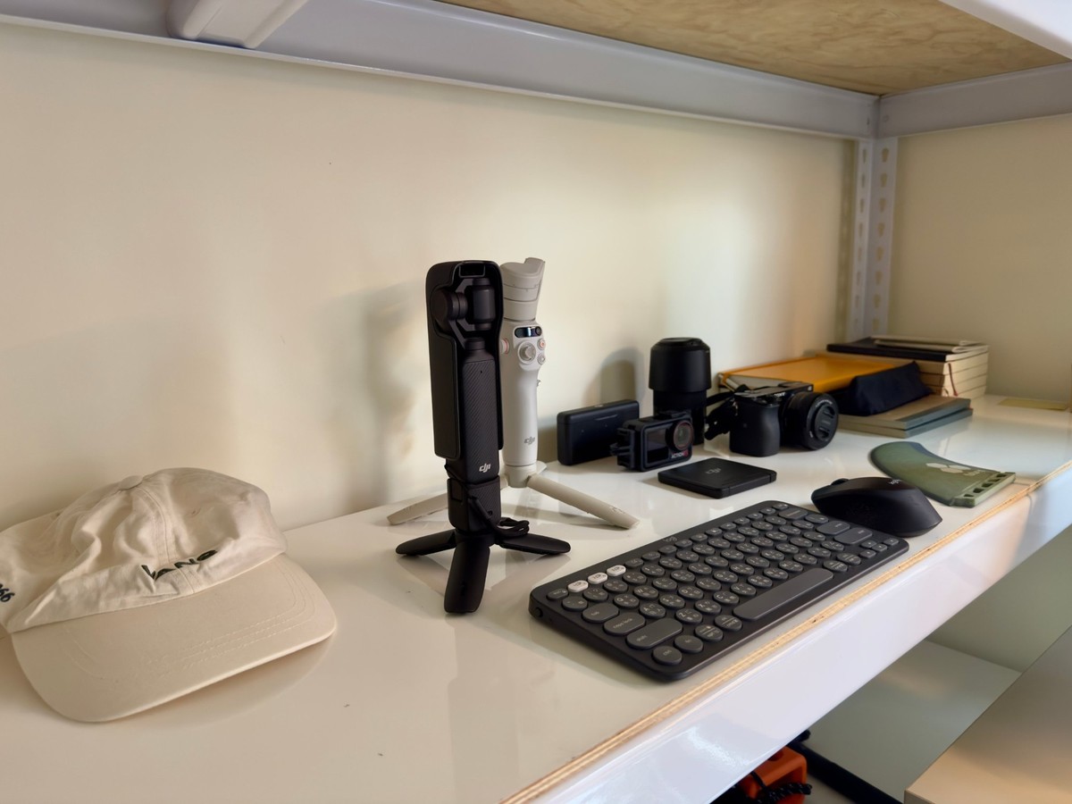 A shelf holding two DJI gimbals on a tabletop tripod, a DJI Osmo Action camera, a Sony mirrorless camera with a lens, a Logitech keyboard and mouse, and a white cap.