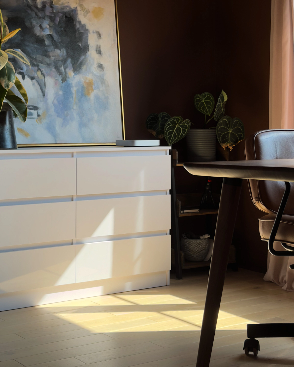 A closer view of the white drawer unit, with the painting above it, anthurium plants on a shelf alongside and the desk leg in the foreground.